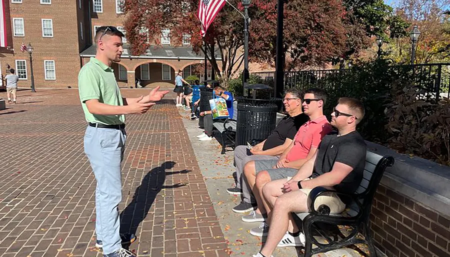 A man in a green shirt is gesturing while speaking to three people seated on a bench in a sunny, outdoor public space.