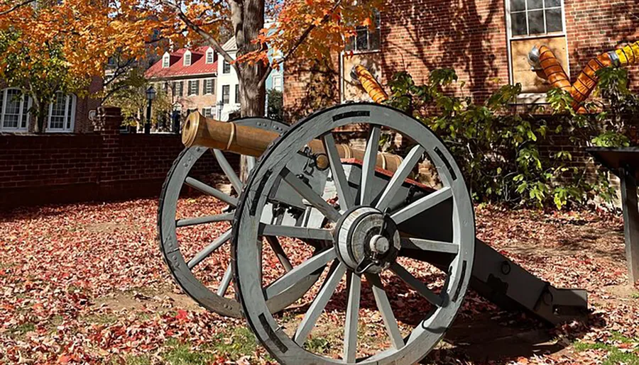 An old-fashioned cannon rests on a carriage amid fallen autumn leaves in a historical setting with colonial-style buildings in the background.