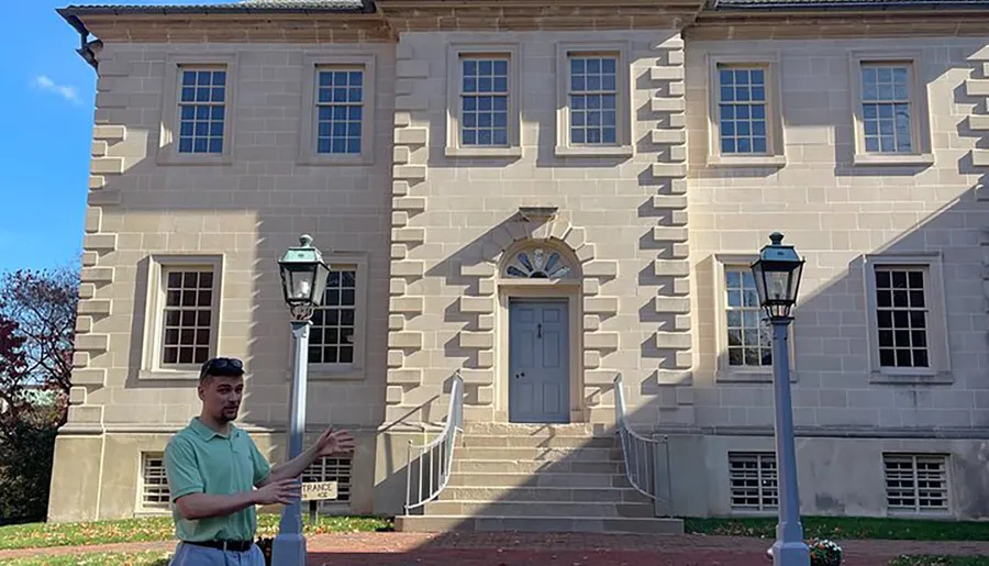 A person is gesturing with their hands while standing in front of a classical building with distinctive quoins and a central doorway.