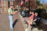 A person is gesturing while speaking to a group of individuals sitting on a bench in a sunny, outdoor public space with American flags in the background.