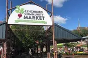 The image shows an entrance sign for the Lynchburg Community Market on a sunny day, with people and market stalls visible in the background.