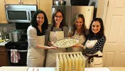 Four smiling women, wearing aprons, are displaying homemade pasta in a kitchen setting.