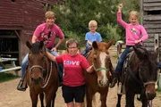 Four people, two of them on horseback and one on a mule, are smiling and posing for a photo at what appears to be a farm or ranch.