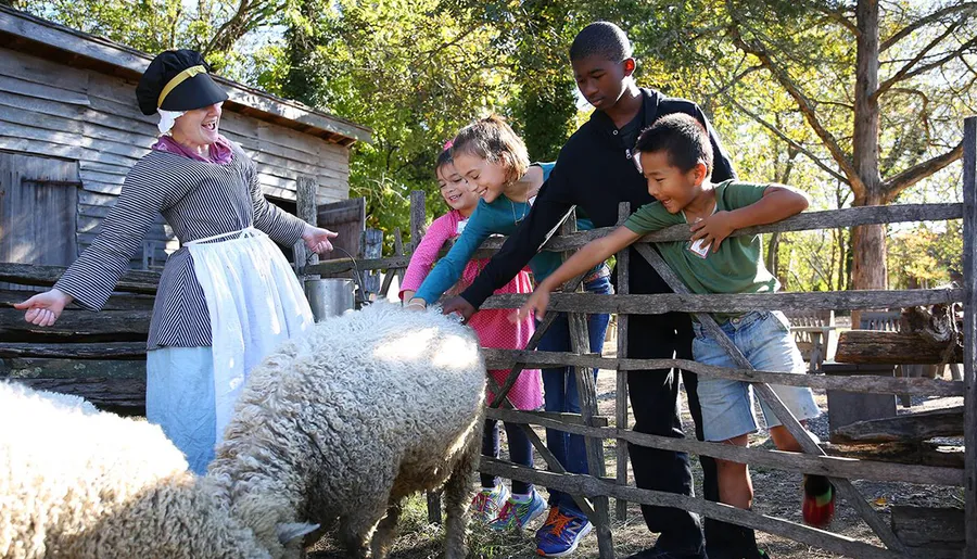 A group of children are petting a sheep at a historical farm under the guidance of a woman dressed in period-appropriate clothing.