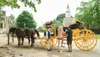 Two people are interacting with a carriage driver dressed in historical attire before a bright yellow horse-drawn carriage, with architectural and natural elements suggesting the scene may be set in a historical or tourist location.