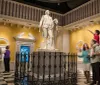 Visitors are interacting with a large statue of a historical figure in a museum-like setting with a child on someones shoulders pointing towards the sculpture