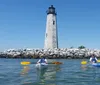 Two people in kayaks are paddling near an old lighthouse surrounded by a rocky breakwater under a clear blue sky