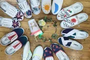 A variety of canvas shoes with different hand-painted designs are arranged in a circle around flowers and a notebook on a wooden floor.