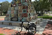 A bicycle with a basket is parked in front of a unique, multi-colored mosaic tower in a sunny outdoor setting.