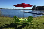 Two green Adirondack chairs and a matching table sit under a red umbrella facing a tranquil lake with a dock and pedal boats.