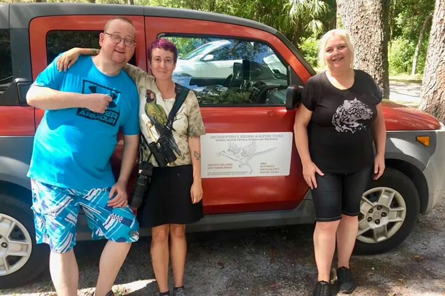 Three people are smiling and posing next to an orange vehicle with a bird watching sign on it.