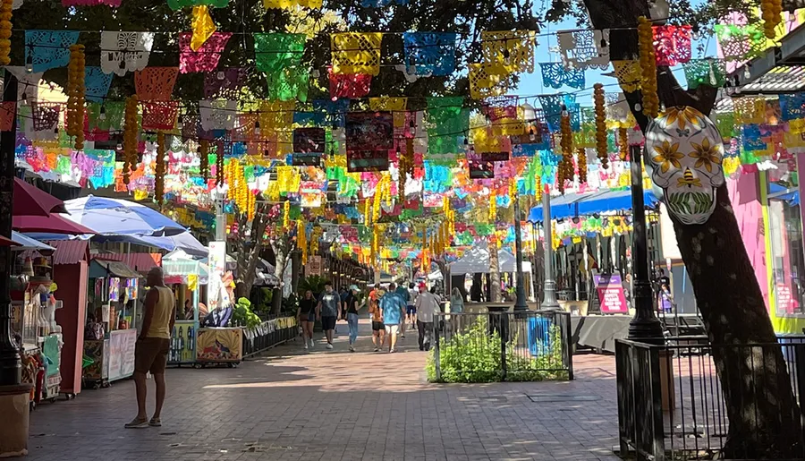 Colorful papel picado banners hang above a bustling outdoor marketplace with pedestrians strolling among vibrant stalls and umbrellas.