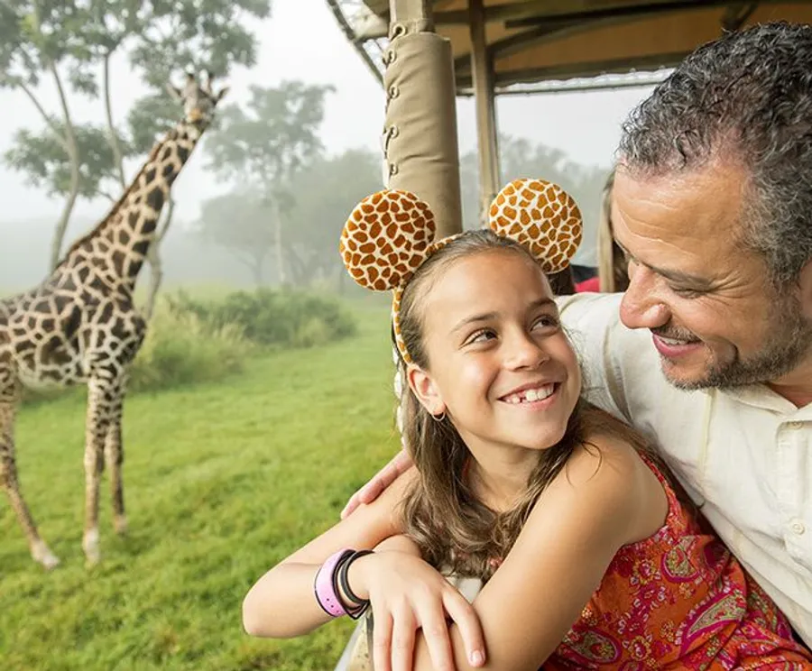 A smiling girl wearing giraffe-ear headband shares a joyful moment with a man, likely her father, while a giraffe stands in the background, possibly at a safari park or zoo.