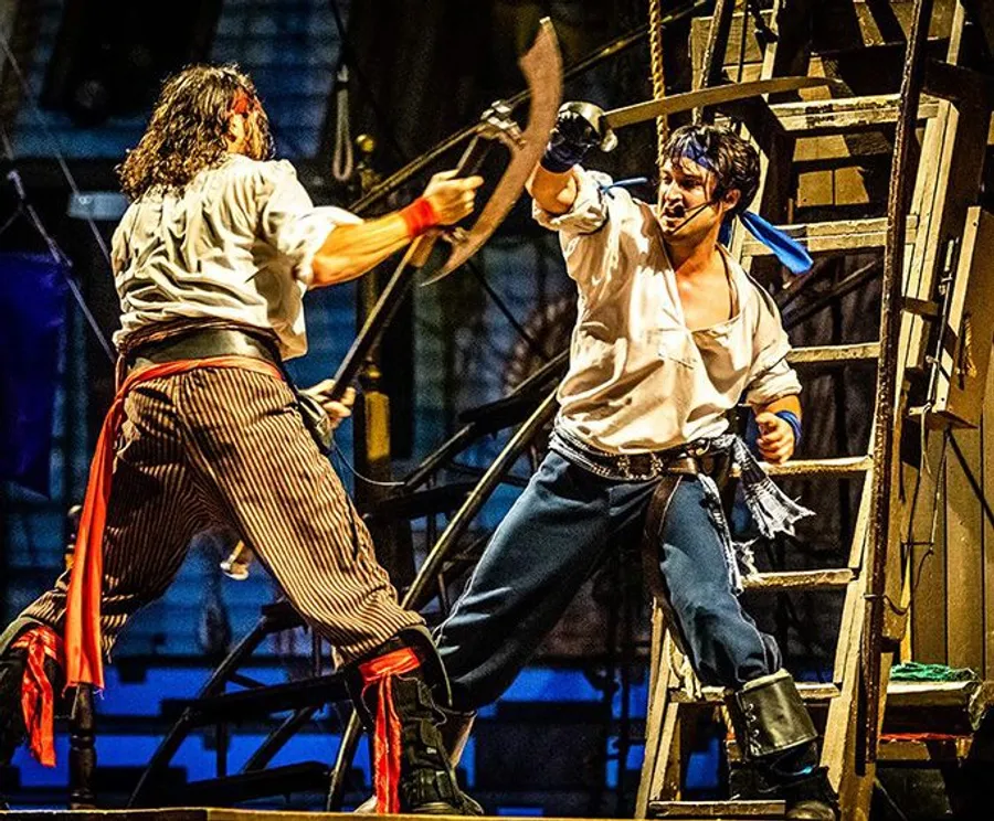 Two actors are engaging in a dramatic swordfight on stage, possibly portraying a scene from a pirate-themed play or performance.