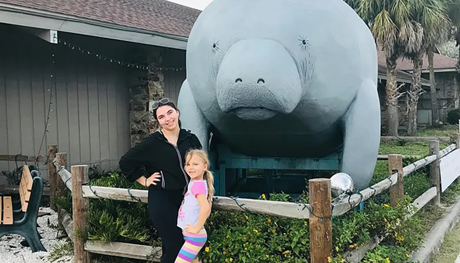 Two people are posing for a photo in front of a large statue of a manatee.