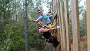 Three people are climbing a tall wooden structure outdoors, secured with harnesses and ropes, displaying expressions of enjoyment and adventure.