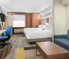 The image shows a modern hotel room with a bed work desk area seating space and abstract wall art combining a functional design with a touch of vibrant colors