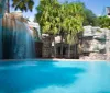 The image shows a serene swimming pool with a cascading artificial waterfall surrounded by tropical vegetation and architectural structures conveying a resort-like atmosphere