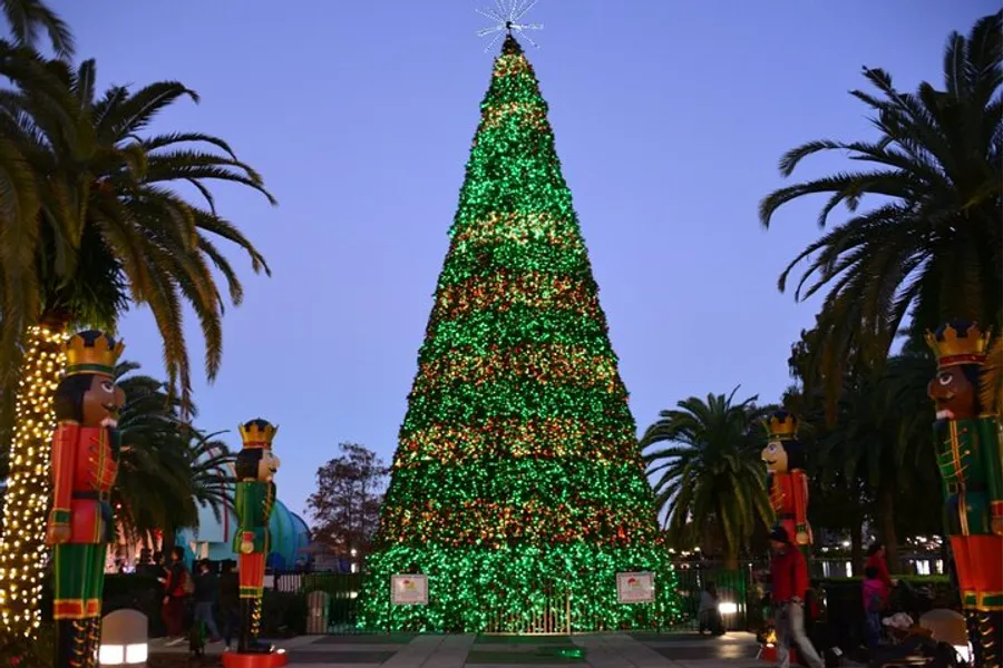 A large, illuminated Christmas tree is adorned with red and green lights, flanked by two nutcracker statues, against a backdrop of twilight and palm trees.