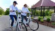 Two individuals are smiling and riding matching white bicycles on a paved path near a gazebo in a park-like setting.