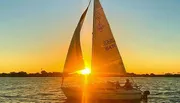 A sailboat glides on the water against a beautiful sunset backdrop, with the sun's rays creating a striking silhouette of the sails.