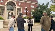 A group of people is standing outside a building, listening to a woman who appears to be giving a tour or presentation.