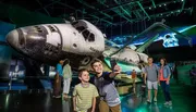 Two boys take a selfie in front of the Space Shuttle Atlantis exhibit while other visitors explore the display.