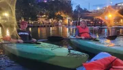 Two individuals are smiling while kayaking in an urban waterway at dusk, with city lights starting to twinkle in the background.