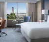 The image shows a modern and neatly arranged hotel room with a large bed a desk seating areas and a window offering a view of a city landscape and a distinctive tower