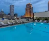 The image shows a tranquil rooftop swimming pool flanked by lounge chairs with a backdrop of the city skyline during dusk