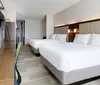 The image shows a modern hotel room with two large beds crisp white bedding minimalist furniture and a bright clean interior
