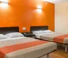 The image shows a simple hotel room with one double bed and two single beds featuring orange walls and minimal furnishings