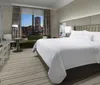 The image shows a neatly arranged hotel room with a large bed modern furnishings and a night view of a cityscape through the window