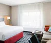 This image shows a well-lit and neatly arranged hotel room with two double beds red accents in the decor a seating area a desk with a television and a large window with sheer curtains