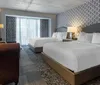 The image shows a modern hotel room furnished with a patterned accent wall a large bed with white bedding matching curtains and a cozy seating area combining contemporary comfort with a touch of classic elegance