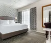 The image shows a modern hotel room furnished with a patterned accent wall a large bed with white bedding matching curtains and a cozy seating area combining contemporary comfort with a touch of classic elegance