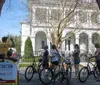 A group of cyclists is stopped to listen to a guide during a bike tour in an urban area with historical houses and lush greenery in the background