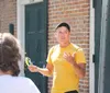 A person in a yellow t-shirt and a cap is talking and gesturing with their hands while holding sunglasses with another persons back to the camera in the foreground and a brick building with a dark green door in the background