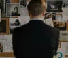 A person is seen from behind contemplating a cork board filled with various photographs notes and strings that suggest a complex investigation or conspiracy