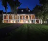 The image depicts a historic two-story plantation-style house with a symmetrical facade illuminated by warm lights at twilight with a lush green lawn in the foreground and draped Spanish moss adding an eerie ambiance