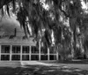 The image depicts a historic two-story plantation-style house with a symmetrical facade illuminated by warm lights at twilight with a lush green lawn in the foreground and draped Spanish moss adding an eerie ambiance