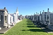 The image shows a sunny day at a cemetery with rows of above-ground tombs and statues, characteristic of a historic or possibly New Orleans-style graveyard, under a clear blue sky.