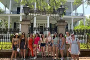 A group of people is posing for a photo in front of a house with a white façade and an ornate wrought iron gate.