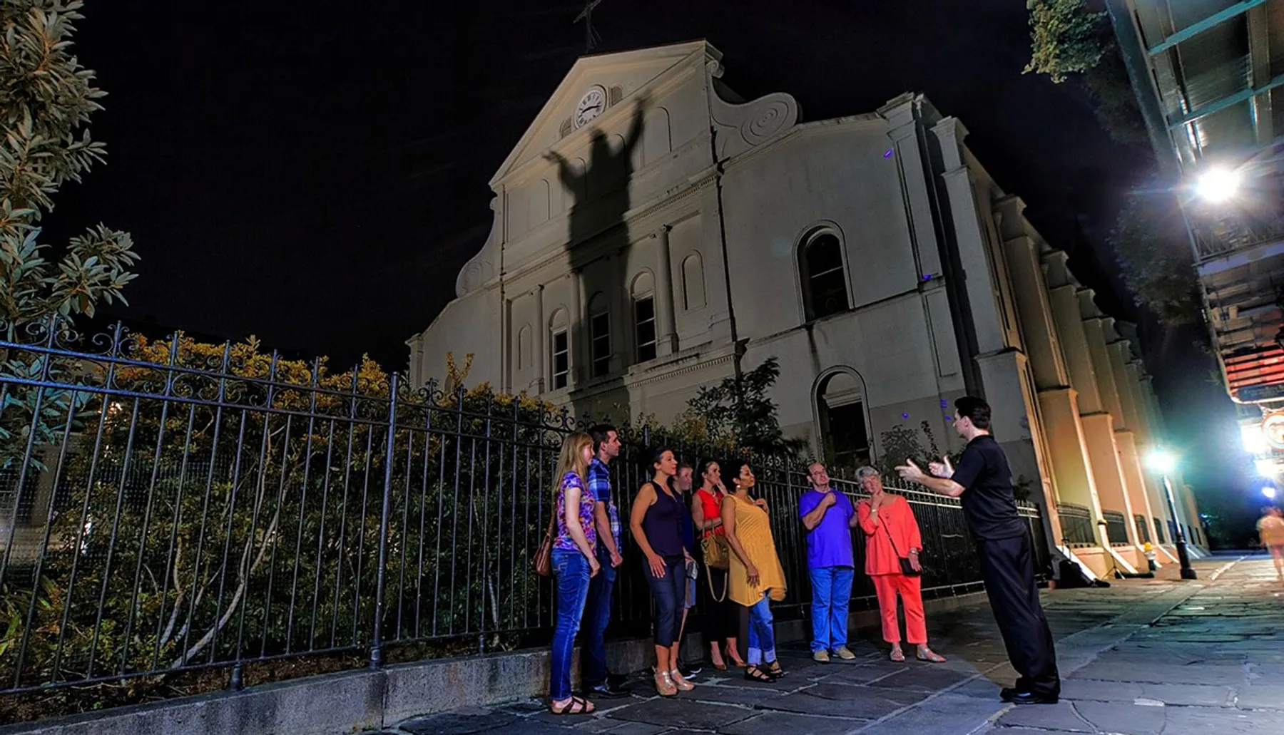 A group of people is attentively listening to a man gesturing while standing in front of a historical building at night.