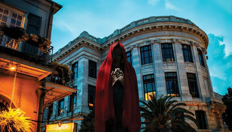 The image shows a large, mysterious figure draped in a red cloak against a backdrop of classical architecture and a twilight sky.