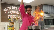 A chef is performing a flambé cooking technique, creating a large flame in a pan, in a kitchen with a sign reading 