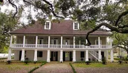 The image shows a two-story white plantation-style house with a covered porch on each level, set among mature oak trees draped with Spanish moss.