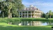A grand plantation-style house with a two-story columned facade overlooks a serene pond with two swans in the foreground amidst lush greenery.