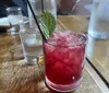 A refreshing crimson-colored cocktail with ice and a mint garnish is served on a wooden table with blurred background of people and other drinks