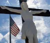 The image shows a statue of Jesus on the cross situated next to an American flag against a backdrop of blue sky and clouds